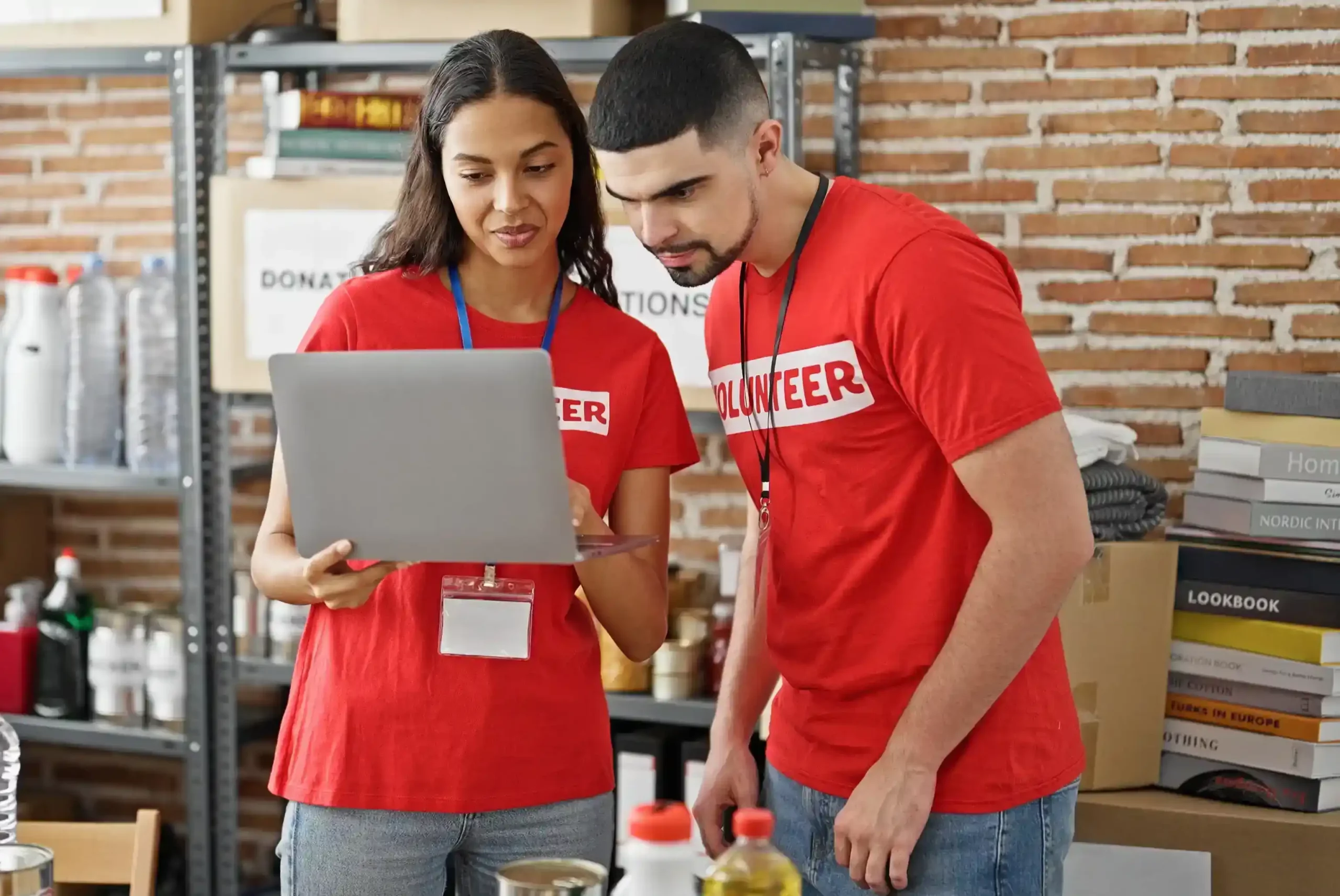Male and female volunteers standing together at a charity center, using a laptop to engage and retain donors. Volunteers collaborating on a laptop at a community center, managing donor updates and personalized communication.
