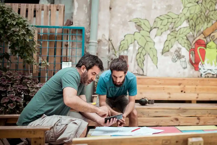 Founders working together in an outdoor workspace and reaching out to Indosoft for advice on customer communication systems. Two startup founders discussing plans while connecting with Indosoft on a smartphone for guidance on scalable communication solutions.
