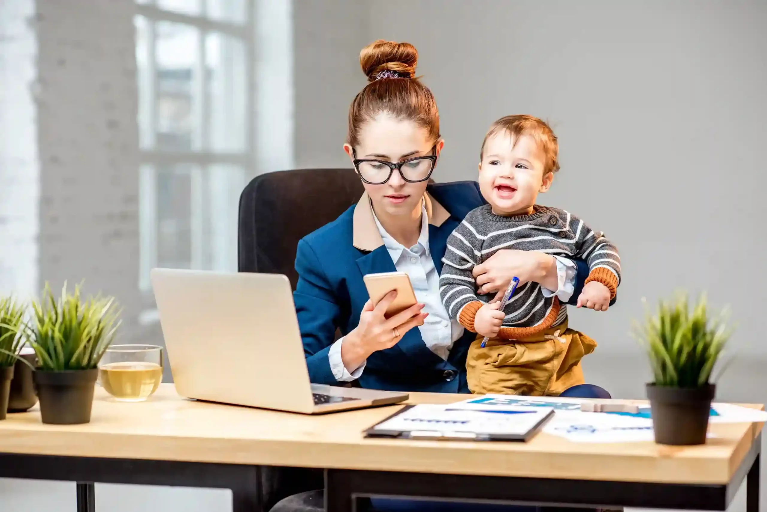 A working mother managing calls and tasks, representing how agents flexibly choose caller IDs that fit different customers and campaigns. A professional parent checking her phone while working at a desk, symbolizing the flexibility to choose the most appropriate calling number for each customer interaction.