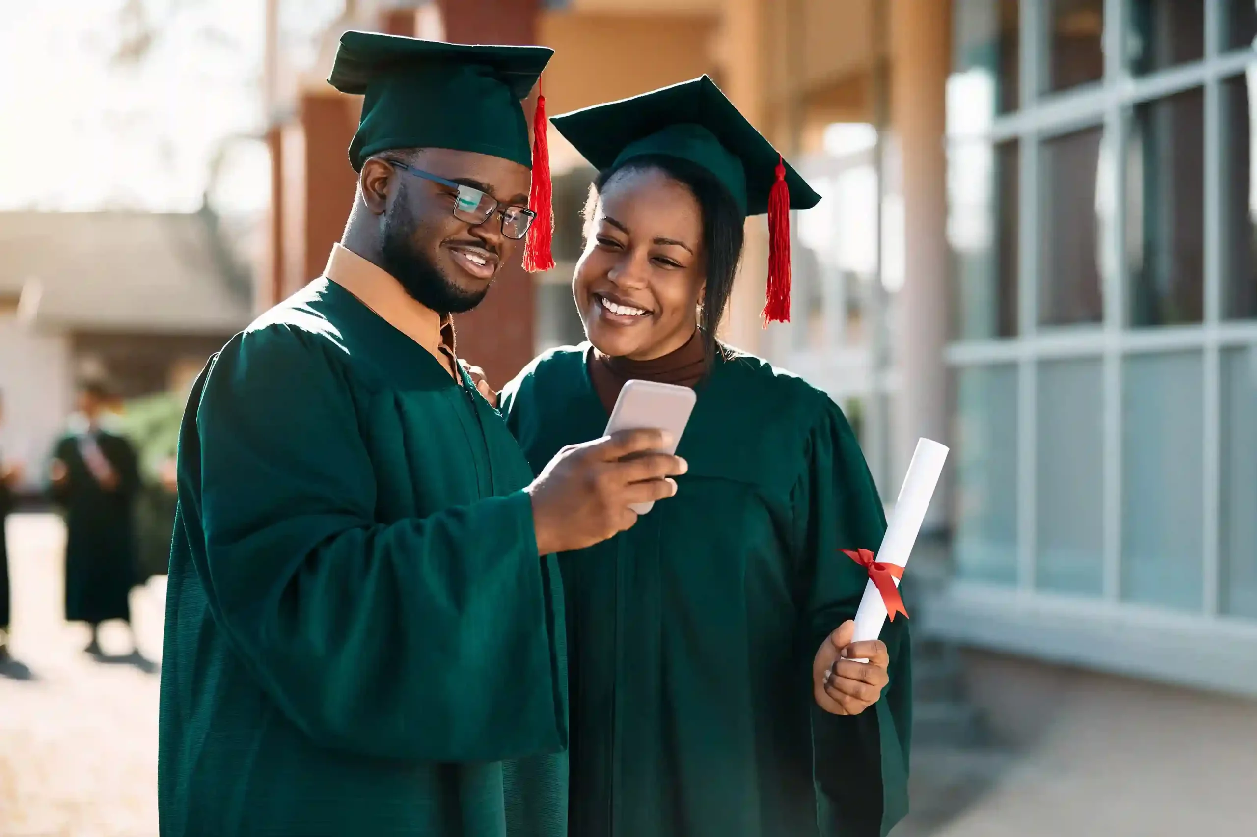 A pair of graduates review a phone message together, symbolizing how standardized greeting messages create a predictable and polished experience for every caller. Two graduates smile while checking a message on a phone, reflecting how every caller receives the same clear and reassuring greeting regardless of timing or team availability.