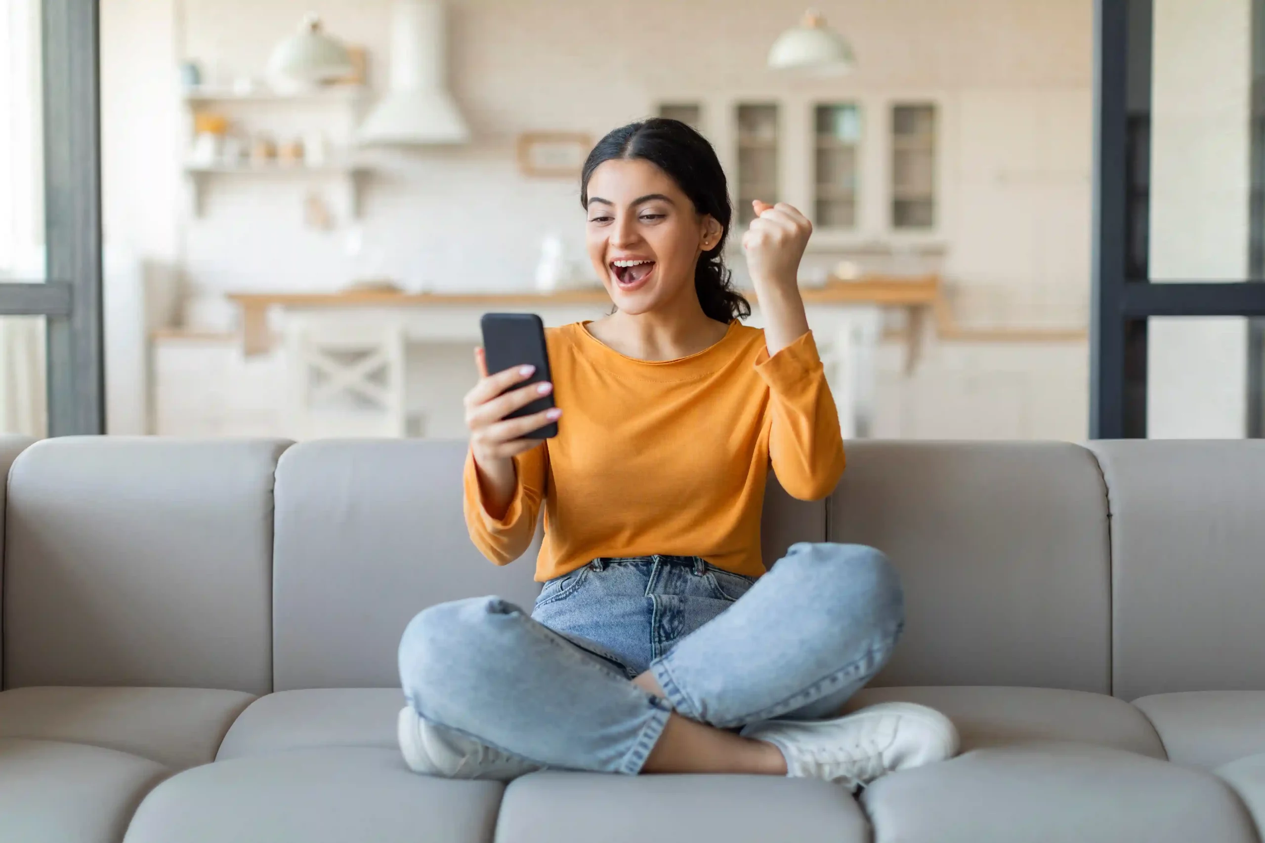 Reacting happily to a game update on her phone, a customer experiences steady SMS delivery that follows predefined filtering rules for consistent communication. Sitting comfortably on her couch, a customer smiles as she receives another timely game update SMS, reflecting how filtered messaging delivers consistent information without confusion.