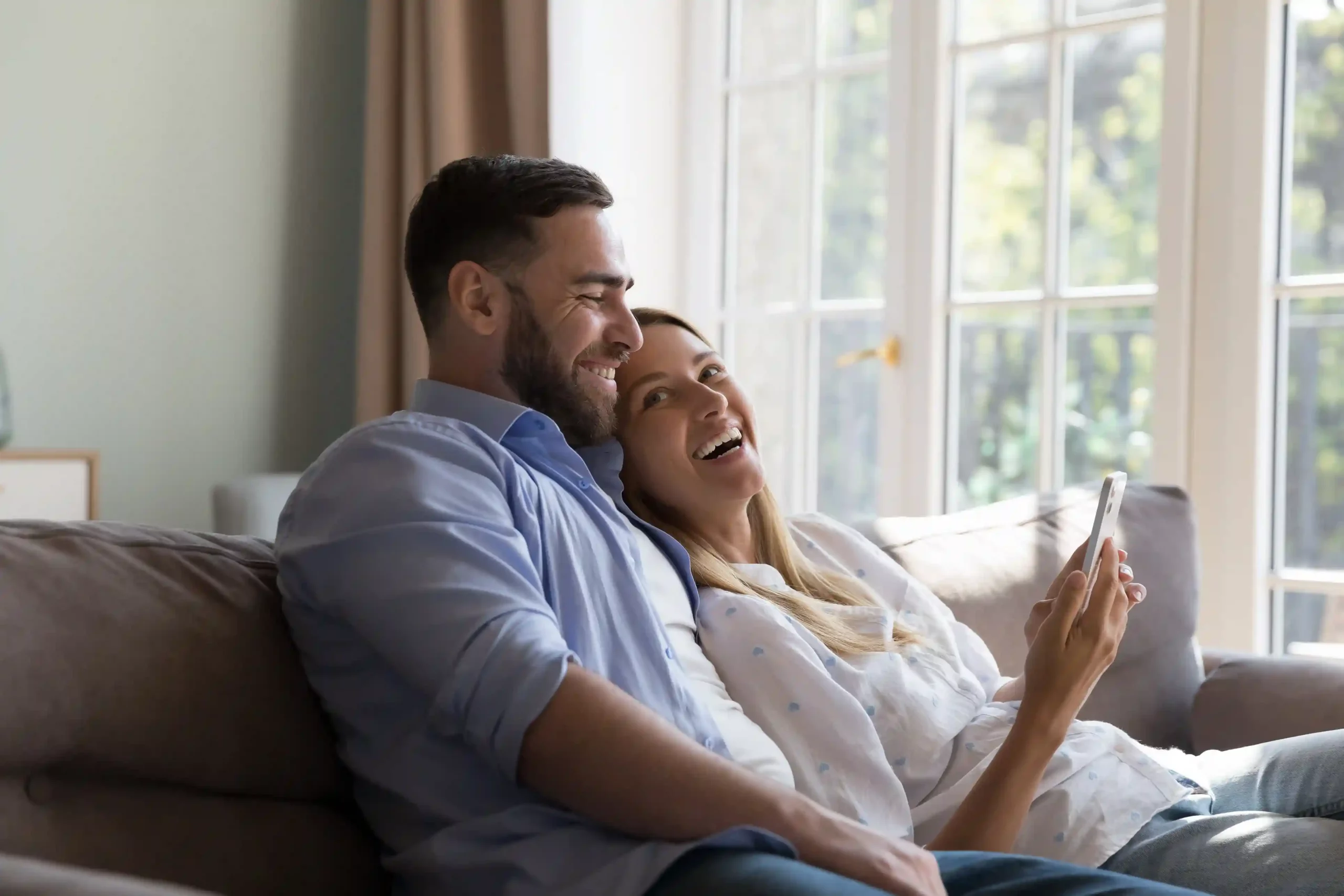 A couple smiles while reviewing details on a phone, benefiting from a well-handled support call where timely guidance kept the situation from escalating. A couple relaxes on their couch after a tax-related call is resolved smoothly, as early agent guidance prevents the issue from escalating further.
