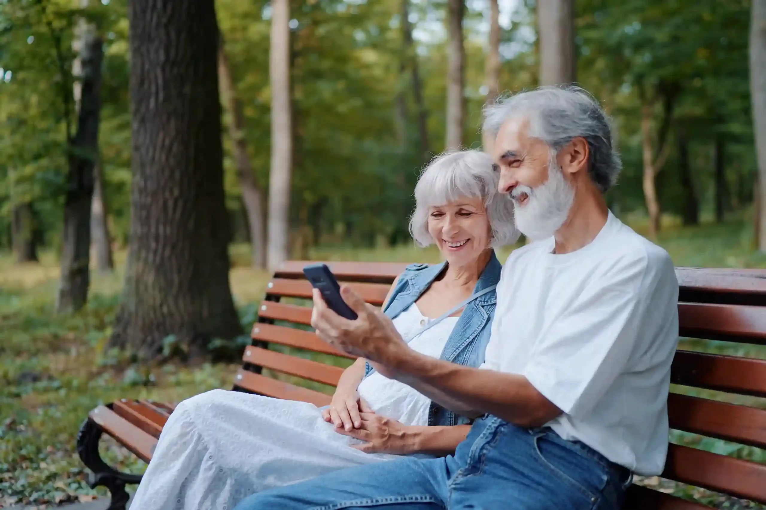 Two seniors smile while looking at a voicemail on a mobile device, illustrating how Indosoft enables voicemail messages to be shared seamlessly with other team members A senior couple sits together on a park bench reviewing a voicemail on a phone, reflecting how Indosoft allows employees to easily forward voicemail messages to teammates for collaboration.