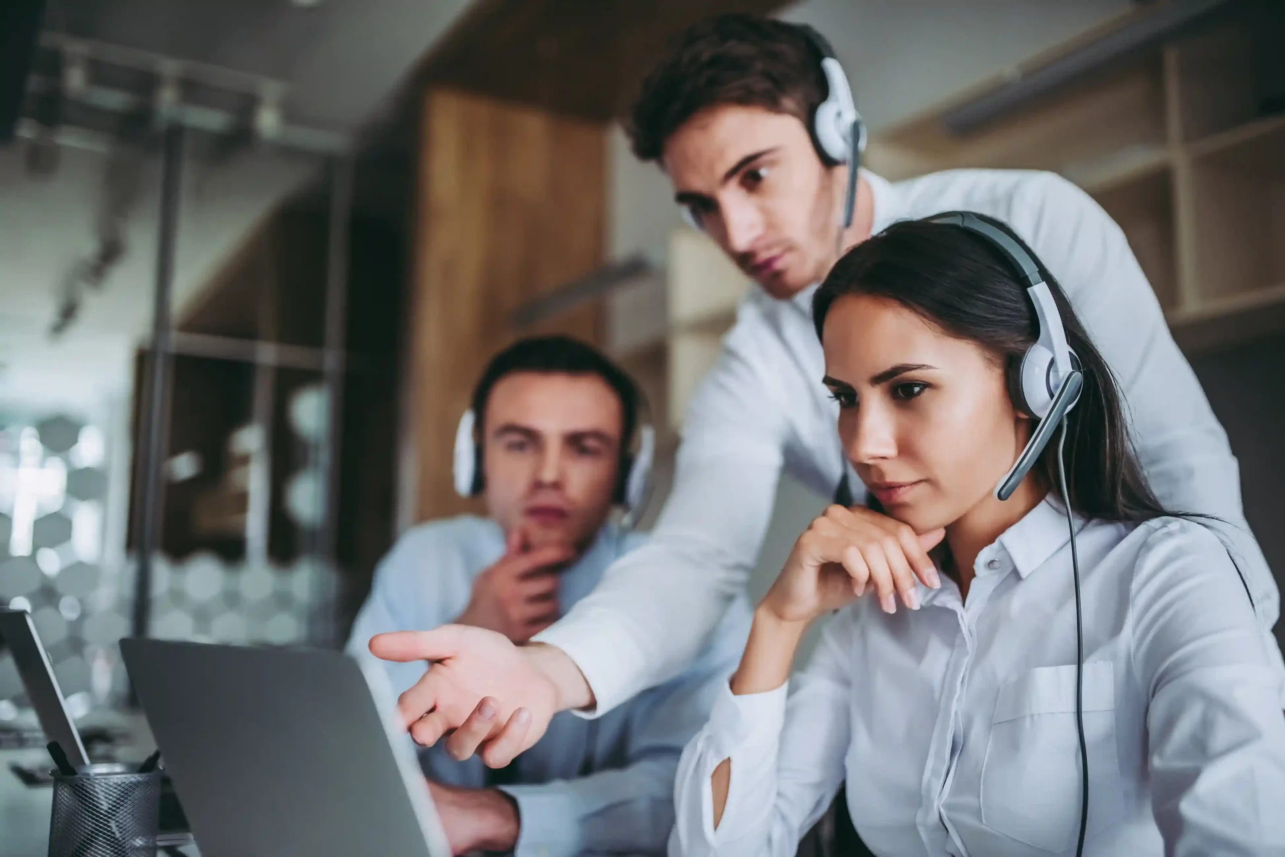 Support agents wearing headsets collaborate over a laptop as smart call filters remove noise from incoming call traffic. A team of contact center agents reviewing call activity together while unwanted call suppression filters out low-value calls before they reach the queue.