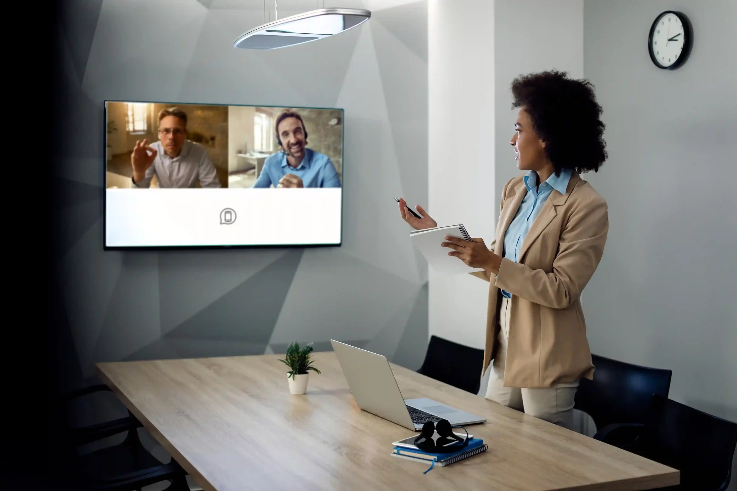 A professional presenter takes notes while speaking to multiple colleagues on a live video screen, reflecting how conference calls support productive discussions across locations. A team leader stands in a meeting room guiding a conference call while remote participants join on a shared screen, showing how Indosoft enables clear, structured group conversations.