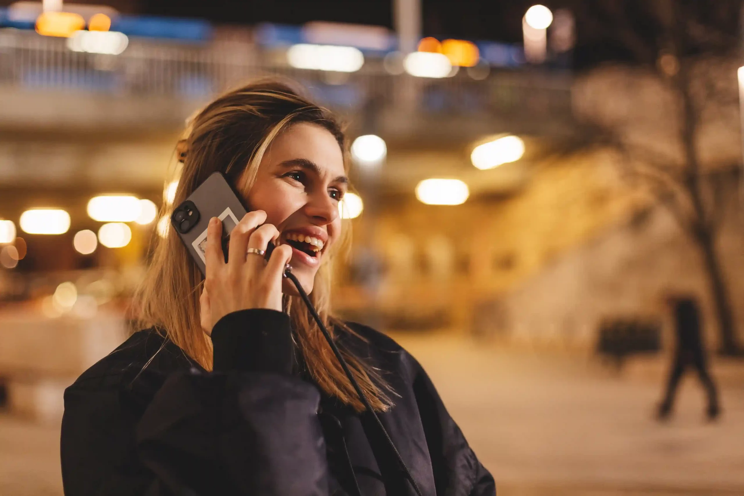 A customer confidently takes a call on her mobile phone outdoors, reflecting how toll-free numbers encourage inbound communication by making it simple and cost-free to reach a business. A woman smiles while talking on her phone in a public space at night, showing how toll-free numbers let customers reach businesses easily without worrying about call costs.