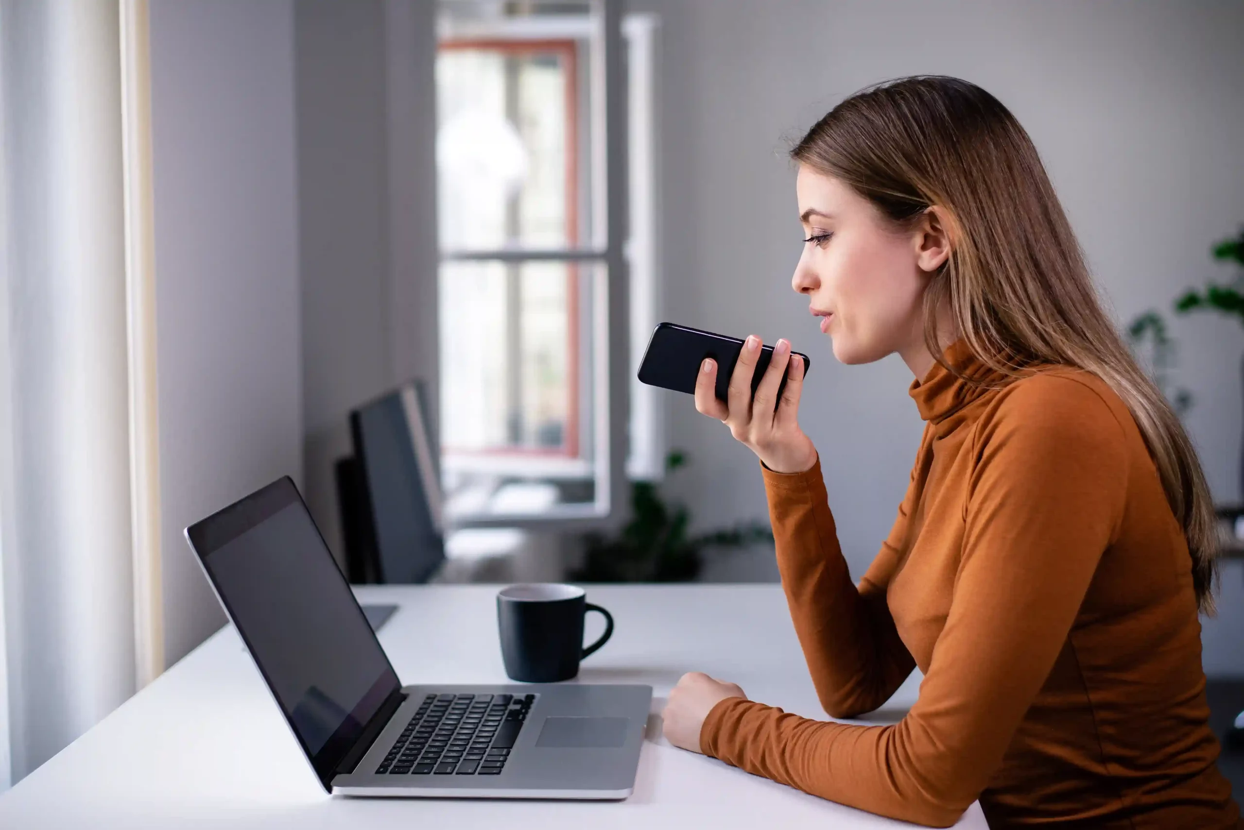 A team member listens to a voicemail on her phone while reviewing information on her laptop, reflecting how voicemail helps businesses track and respond to missed calls efficiently. A professional records a voice message on her phone while working at her desk, showing how voicemail captures important customer messages when live calls cannot be answered.
